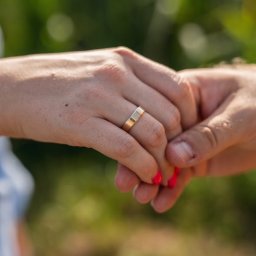 Namika Media Oliwia Nowicka - Dłonie pary trzymające się na tle zieleni, z widocznym złotym pierścionkiem na palcu kobiety z czerwonymi paznokciami. Delikatne światło podkreśla detale.
