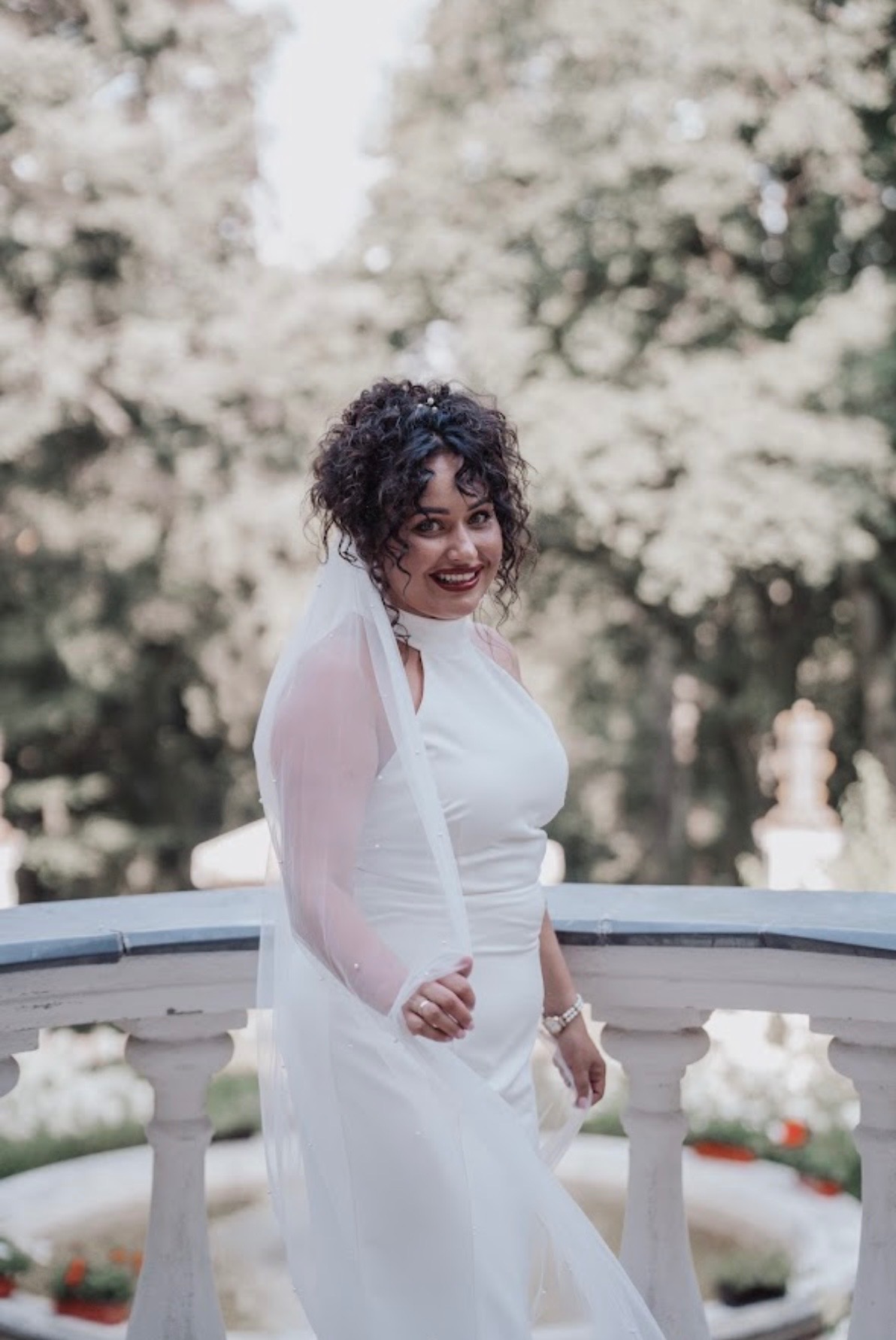 Uśmiechnięta panna młoda w białej sukni i welonie, pozująca na tle parku z balustradą. Delikatne światło podkreśla kręcone włosy i makijaż.