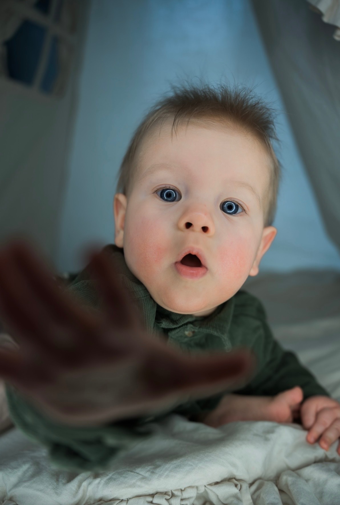 Niemowlę z szeroko otwartymi oczami i ustami wyciąga rączkę w stronę obiektywu, ubrane w zieloną koszulę, leżące na białym tle w namiocie tipi.