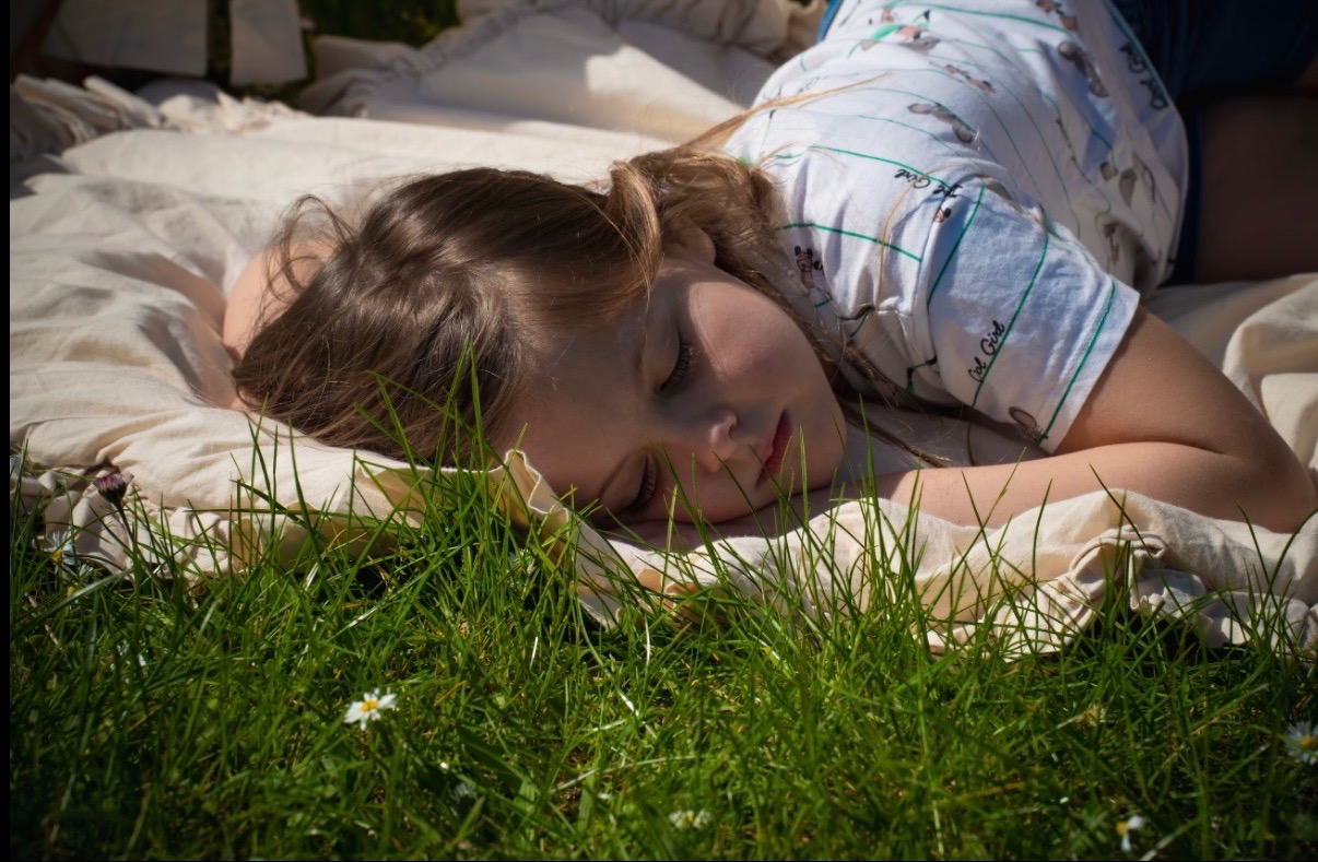 Śpiąca dziewczynka w koszulce z nadrukiem leży na kocu na trawie, z bliska, w naturalnym świetle. Ujęcie z poziomu trawy.