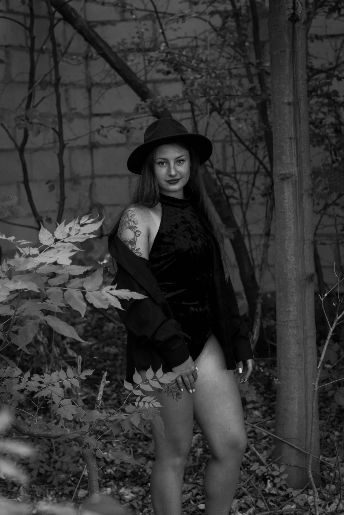Czarno-białe zdjęcie kobiety w czarnym body i kapeluszu, pozującej w plenerze z roślinnością. Widoczny tatuaż na ramieniu i delikatny uśmiech.