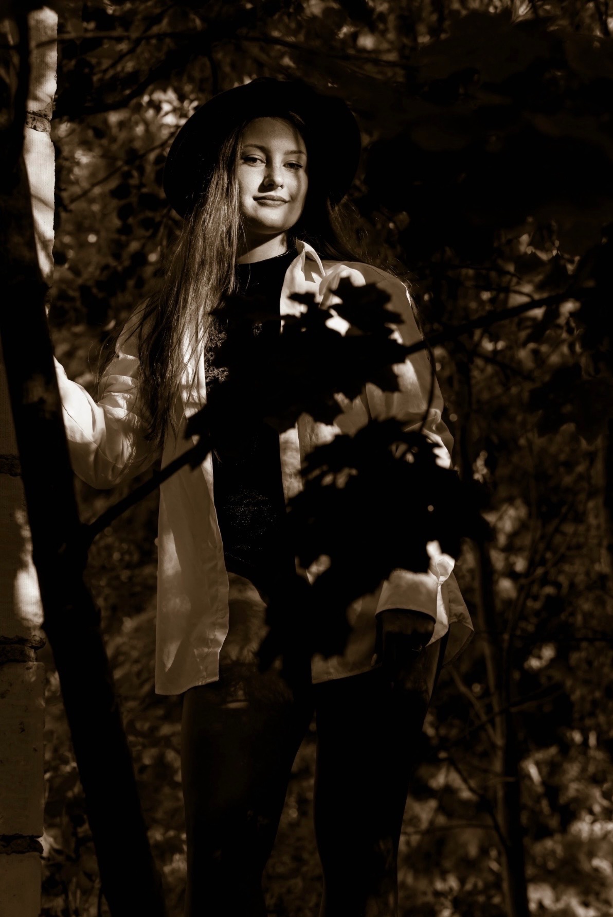 Portret kobiety w kapeluszu, w leśnej scenerii, w sepii. Ubrana w białą koszulę i czarny top, opierająca się o drzewo. Ciemne liście w tle tworzą głębię.