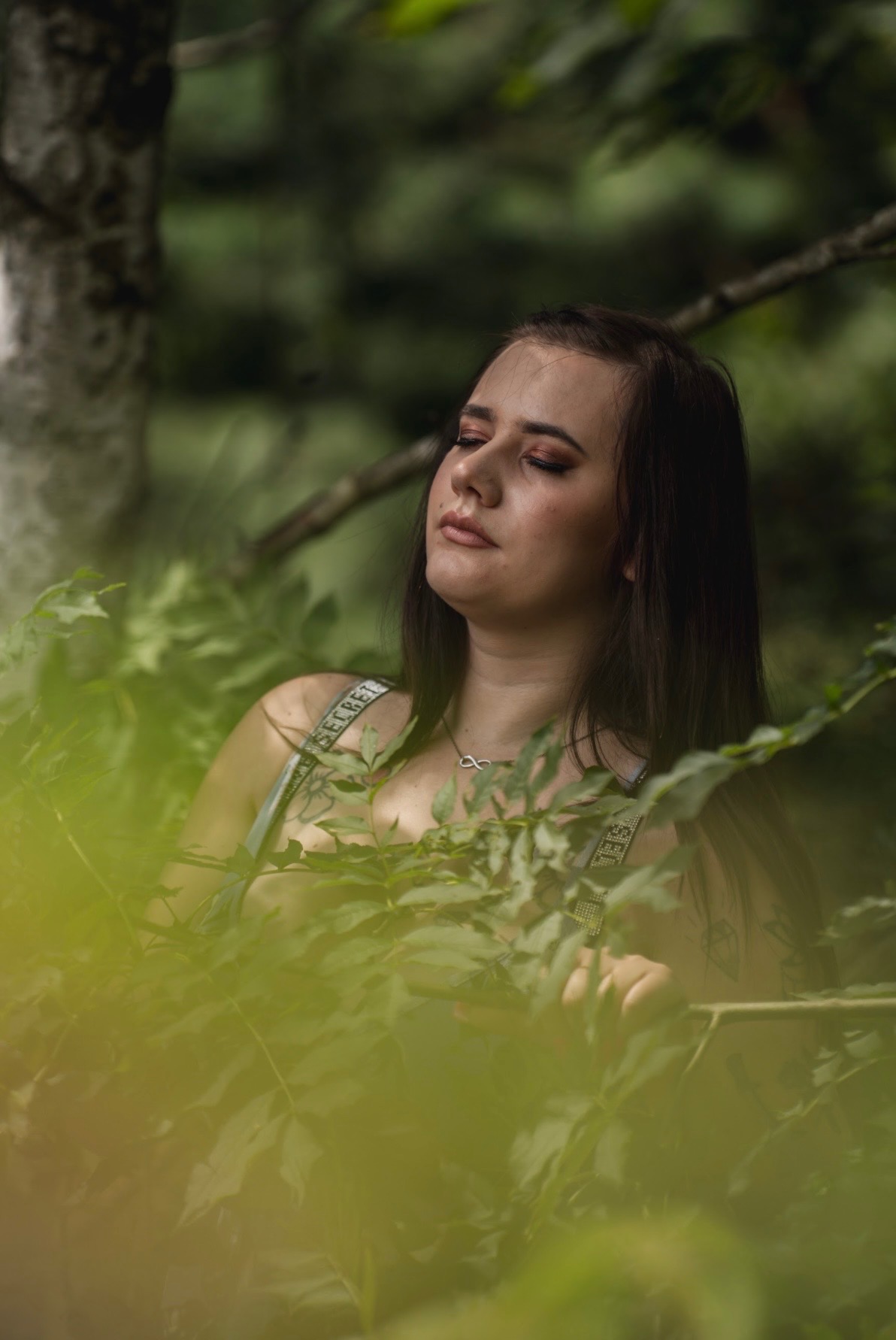 Portret kobiety z zamkniętymi oczami, otoczonej zielenią, w naturalnym oświetleniu. Ujęcie z dołu, podkreślające subtelność i spokój.