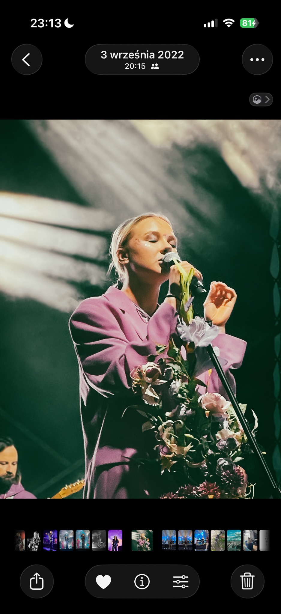 Piosenkarka w różowym garniturze śpiewa do mikrofonu ozdobionego kwiatami na scenie oświetlonej reflektorami. W tle widoczny muzyk z gitarą.