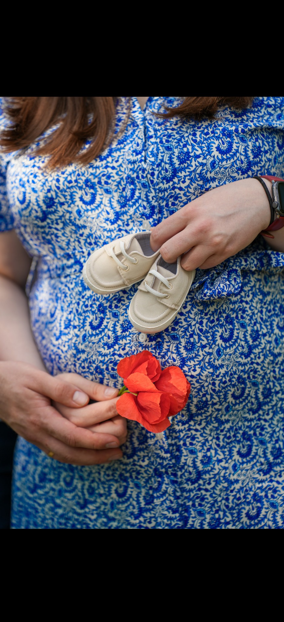 Brzuch kobiety w kwiecistej sukience, trzymającej małe buciki i czerwony mak. Ujęcie z sesji ciążowej, skupiające się na symbolice oczekiwania na dziecko.