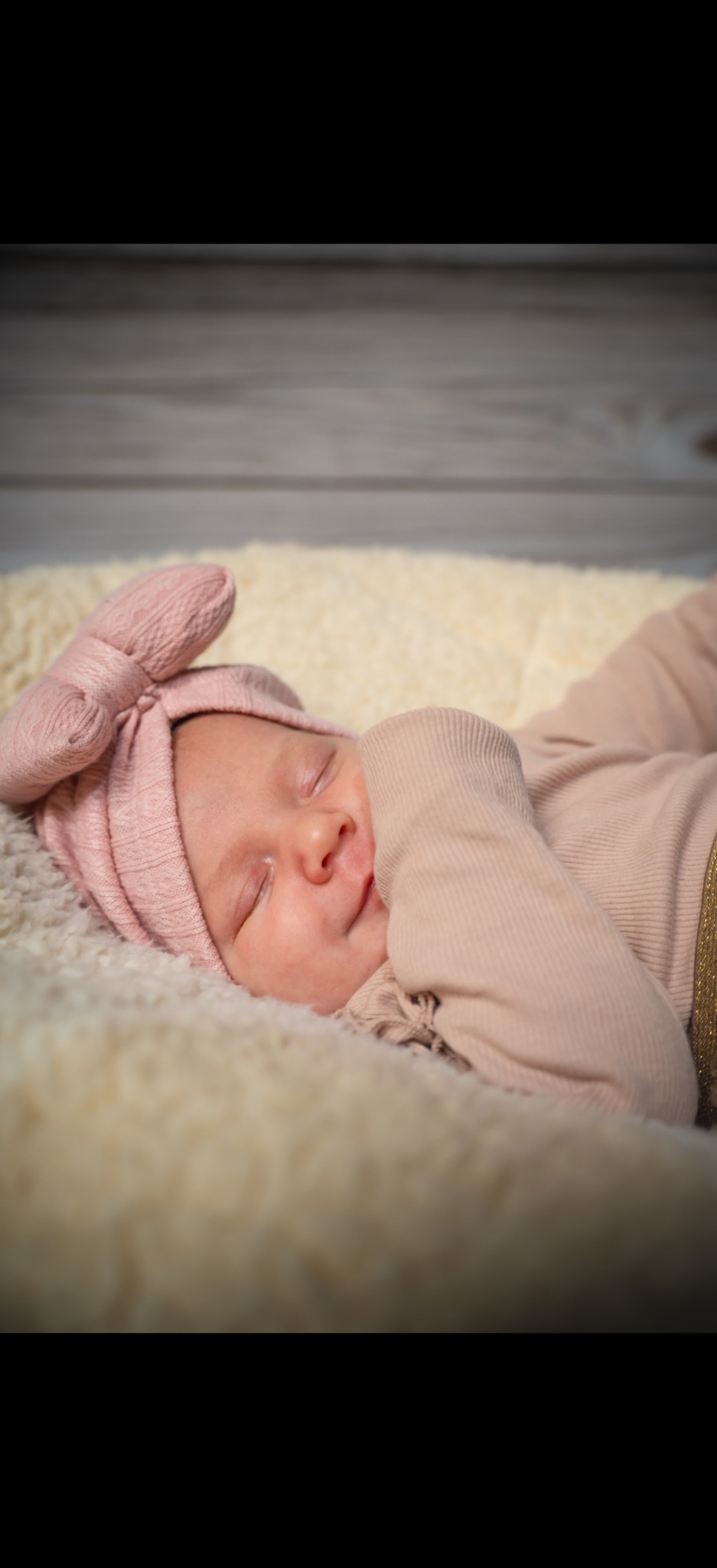 Śpiąca noworodka w różowym ubranku i opasce z kokardą, leżąca na puszystym, kremowym kocyku na tle drewnianej podłogi. Delikatne światło podkreśla rysy twarzy.