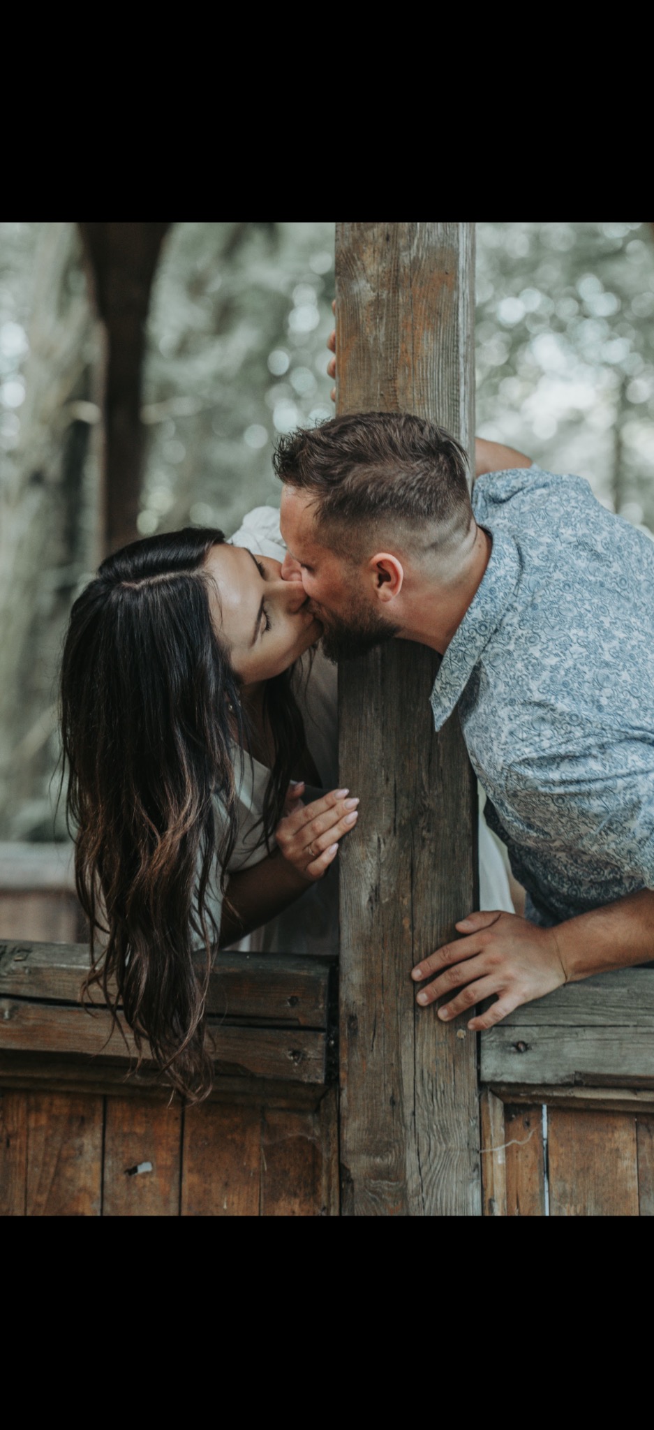 Zakochana para całuje się, opierając się o drewnianą konstrukcję na tle zieleni. Kadr z sesji narzeczeńskiej, delikatne światło, naturalna sceneria.
