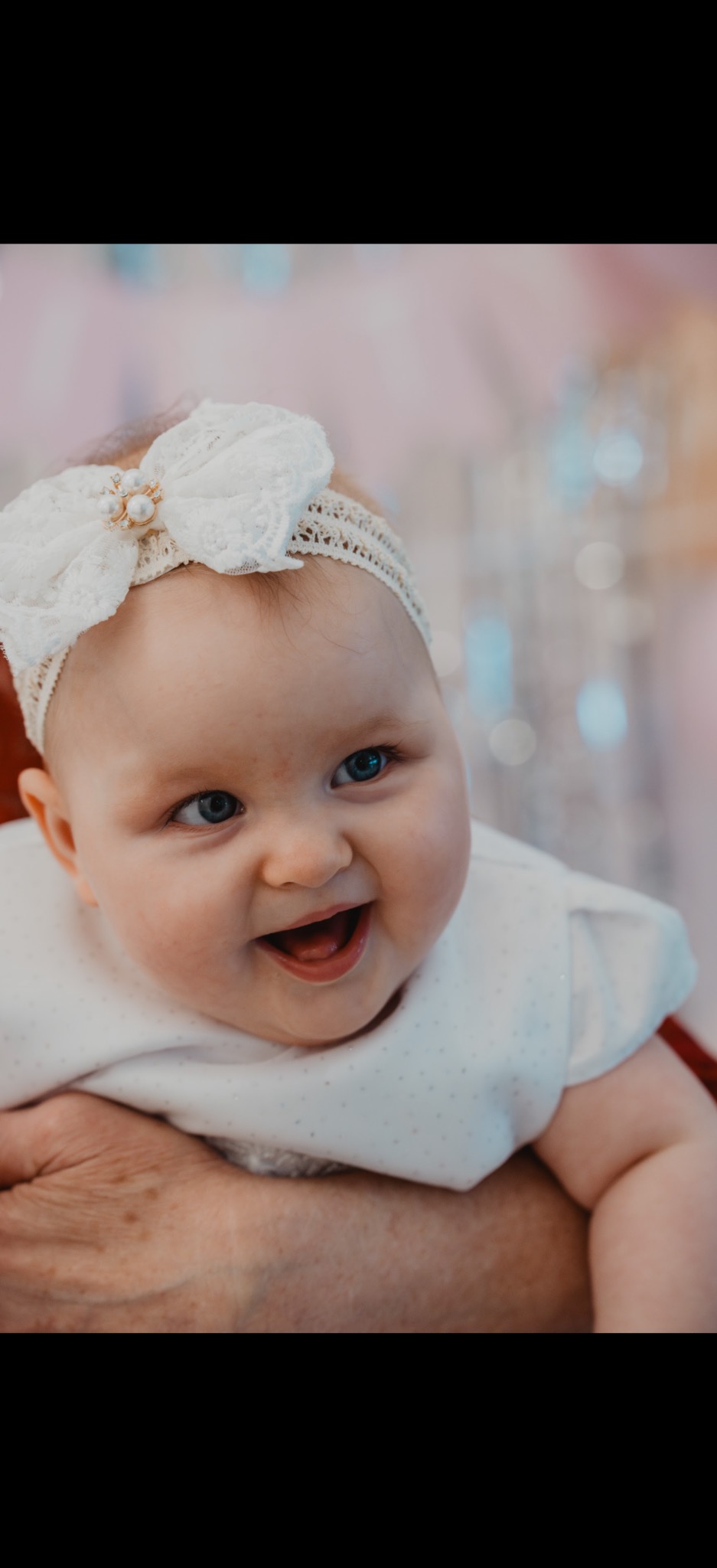 Uśmiechnięta niemowlęca dziewczynka z niebieskimi oczami i białą koronkową opaską na głowie, trzymana na rękach, w tle rozmyte dekoracje.