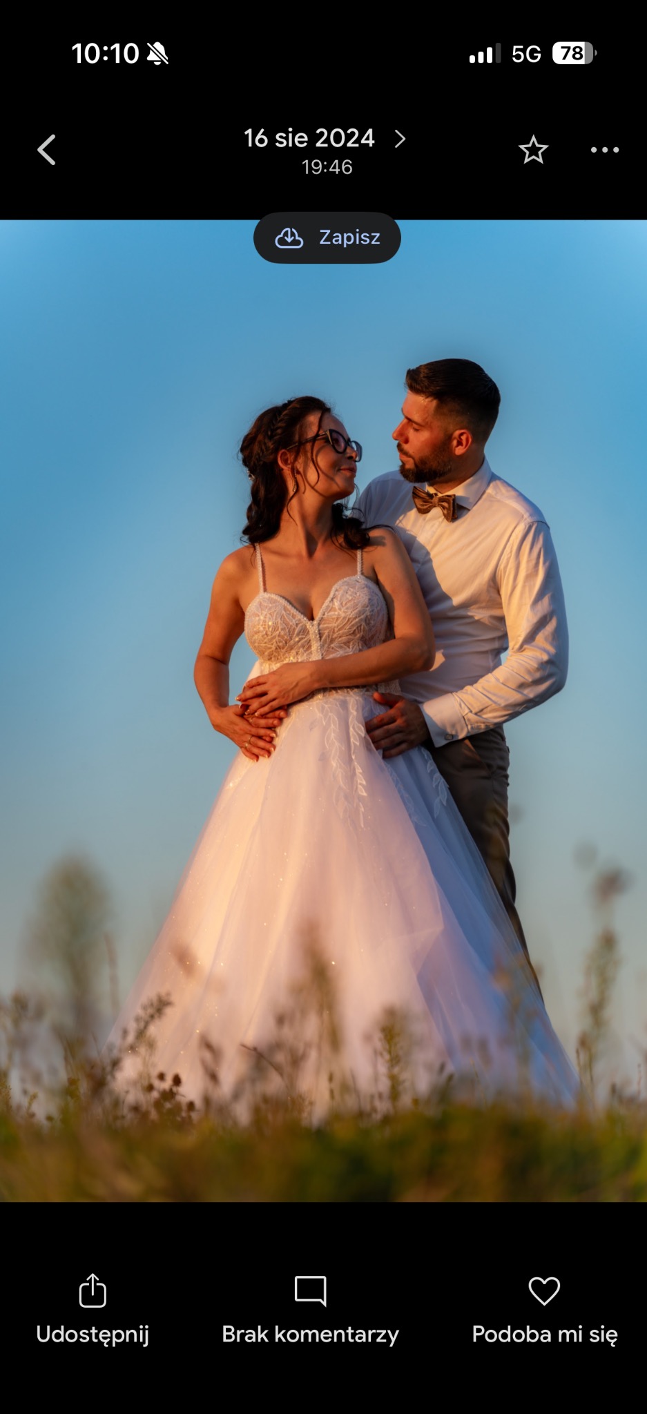 Para młoda w objęciach na tle błękitnego nieba i łąki. Panna młoda w okularach i sukni z koronkową górą, pan młody w muszce. Romantyczna sesja w plenerze.