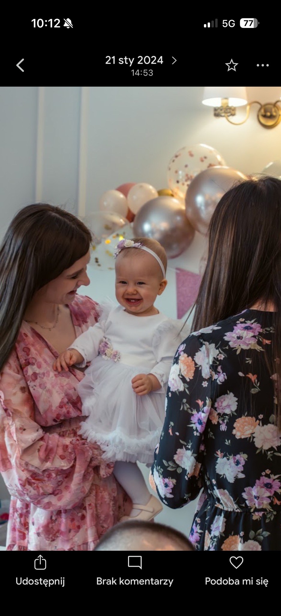 Uśmiechnięta dziewczynka w białej sukience na tle balonów, trzymana przez kobietę w Bogdanowie. Radosna atmosfera, jasne oświetlenie, dekoracje urodzinowe.