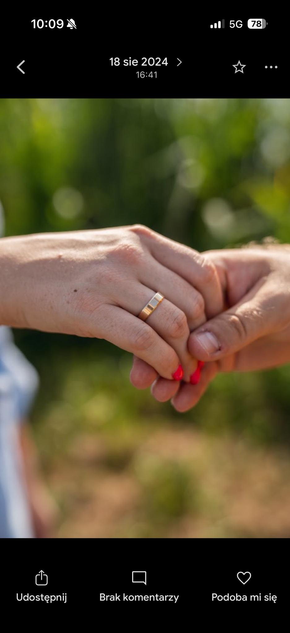 Dłonie pary trzymające się na tle zieleni, z widocznym złotym pierścionkiem na palcu kobiety z czerwonymi paznokciami. Delikatne światło podkreśla detale.