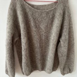 Olesia Liutkevych - Zdjęcie beżowego, ręcznie robionego swetra z włóczki moherowej, wiszącego na czerwonym wieszaku na tle białej ściany. Widoczna faktura dzianiny i luźny krój.