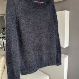 Olesia Liutkevych - Czarny, wełniany sweter z długim rękawem, wiszący na różowym wieszaku na tle białych półek i szarej ściany. Widoczny fragment sofy z jasnymi poduszkami.