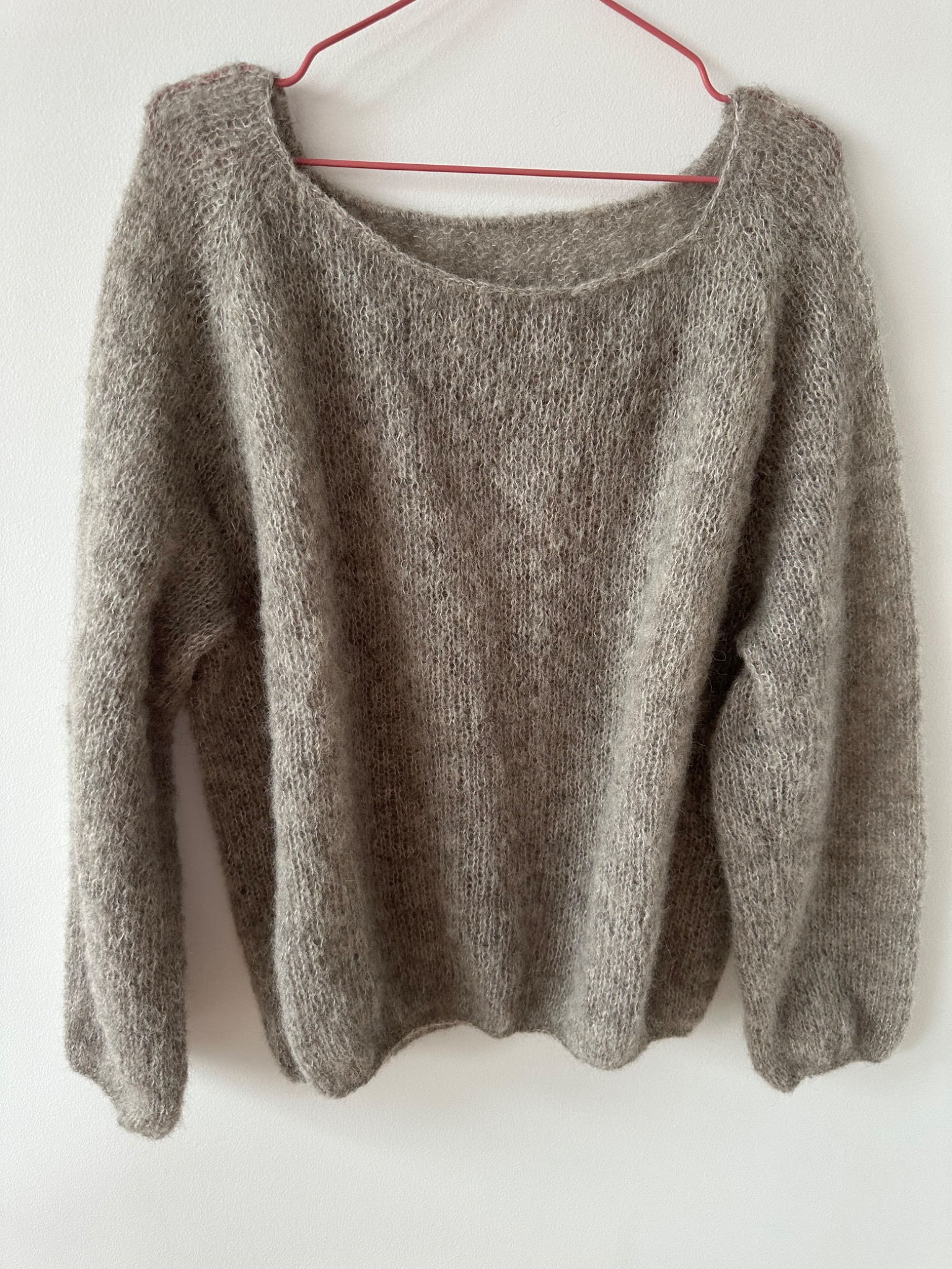 Zdjęcie beżowego, ręcznie robionego swetra z włóczki moherowej, wiszącego na czerwonym wieszaku na tle białej ściany. Widoczna faktura dzianiny i luźny krój.