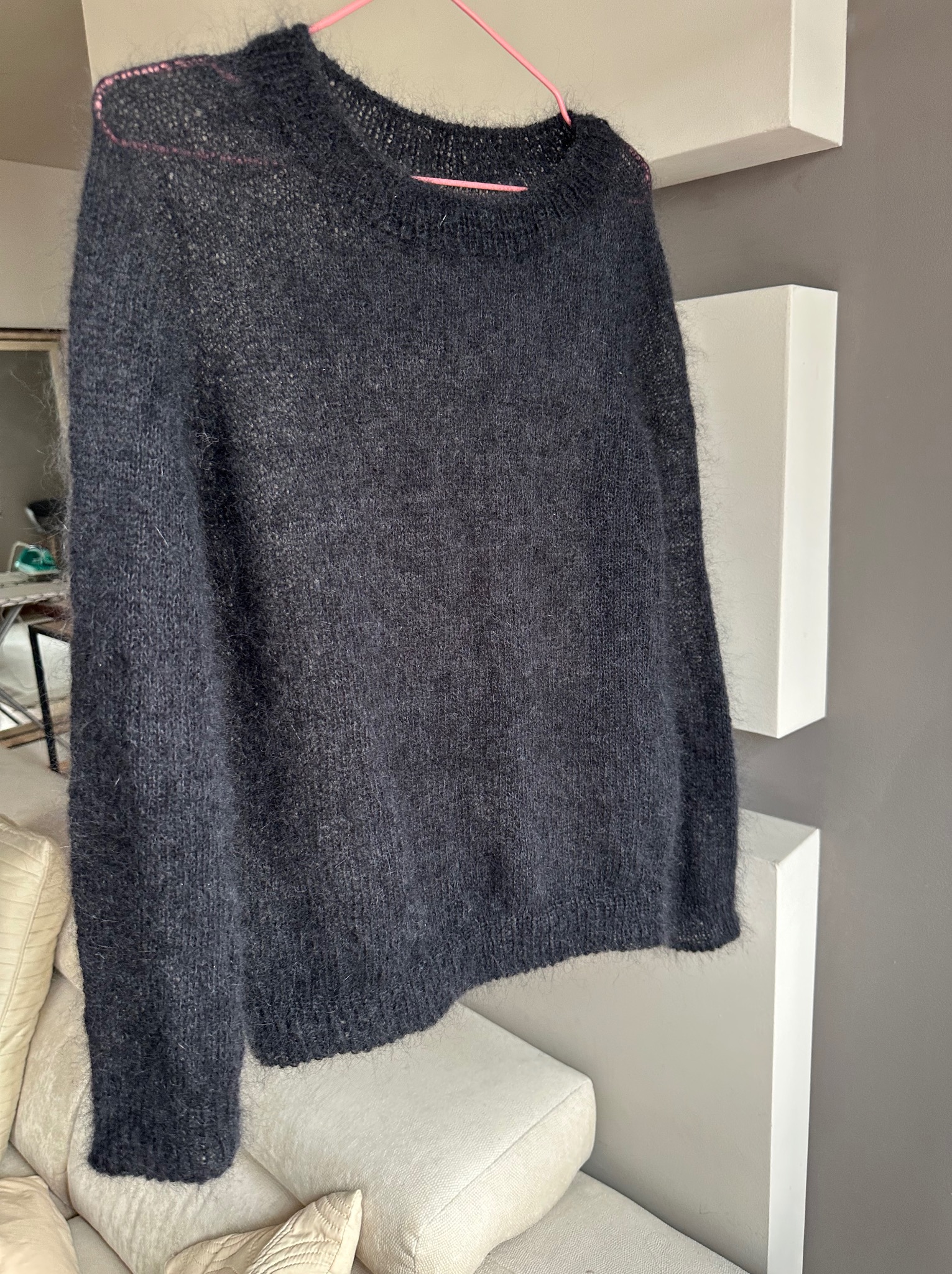 Czarny, wełniany sweter z długim rękawem, wiszący na różowym wieszaku na tle białych półek i szarej ściany. Widoczny fragment sofy z jasnymi poduszkami.