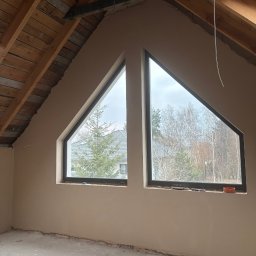 Perfect Tynk Albert Barcikowski - Poddasze po tynkowaniu maszynowym: Dwa trójkątne okna z widokiem na drzewa. Ściany w jasnym odcieniu, surowy beton na podłodze. Widoczne drewniane elementy konstrukcji dachu.