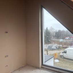 Perfect Tynk Albert Barcikowski - Wnętrze poddasza z oknem o nietypowym kształcie, ściany po nałożeniu gładzi, widoczne puszki elektryczne. Na zewnątrz widok na plac budowy i zaparkowany biały bus.