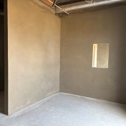 Perfect Tynk Albert Barcikowski - Wnętrze po tynkowaniu maszynowym: gładkie, beżowe ściany i szara posadzka. Widoczne instalacje pod sufitem i otwór okienny. Minimalistyczny kadr, surowy stan wykończenia.