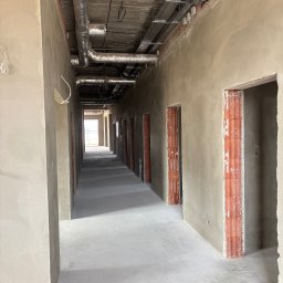 Perfect Tynk Albert Barcikowski - Długi korytarz w surowym stanie po tynkowaniu maszynowym, widoczne instalacje pod sufitem i ceglane obramowania drzwi. Świeże tynki na ścianach i posadzce.