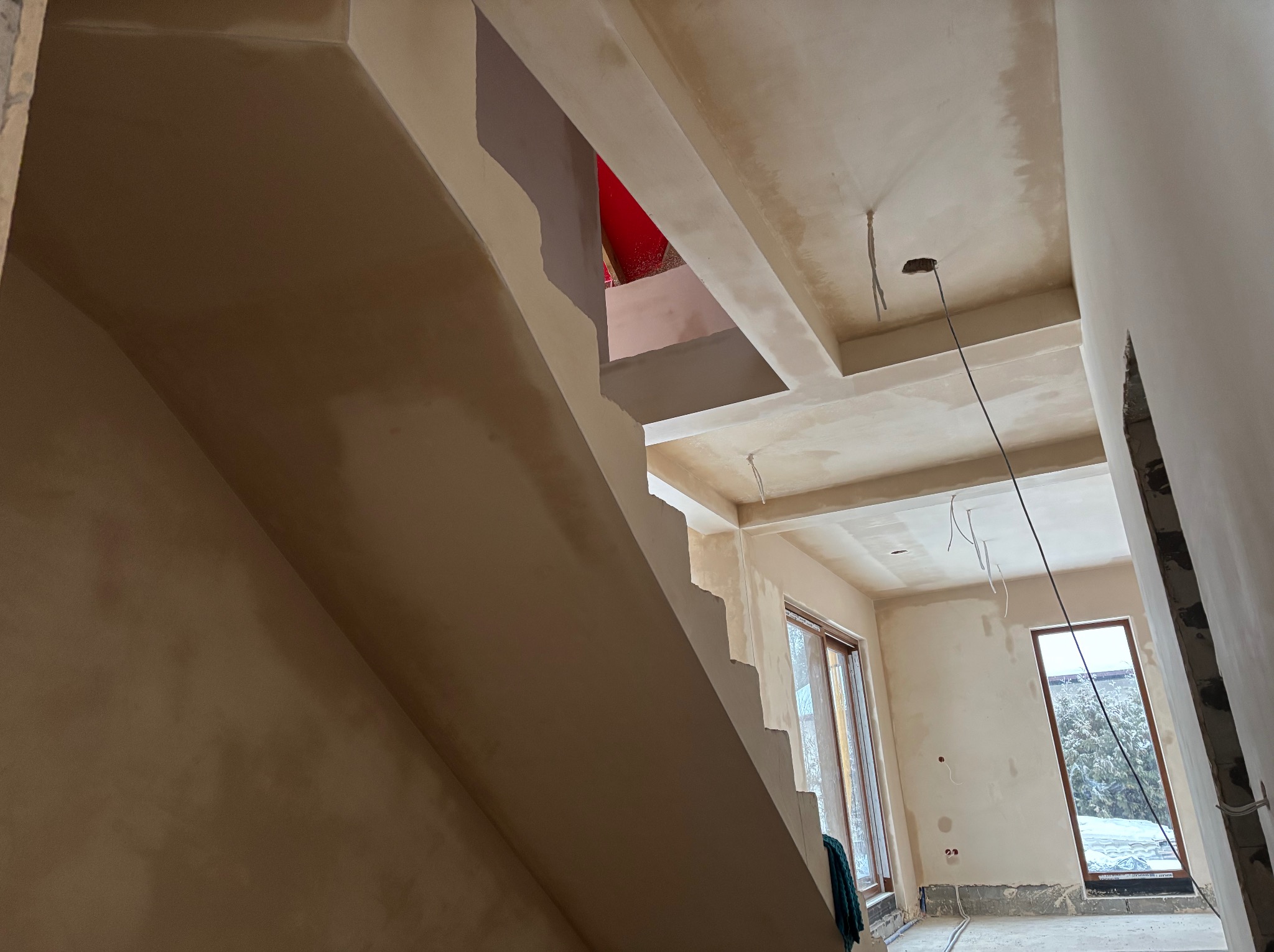 Wnętrze domu w trakcie prac wykończeniowych: otynkowane ściany i sufit, widoczne schody i okna. Przewody elektryczne wystają z sufitu, co wskazuje na etap instalacji.