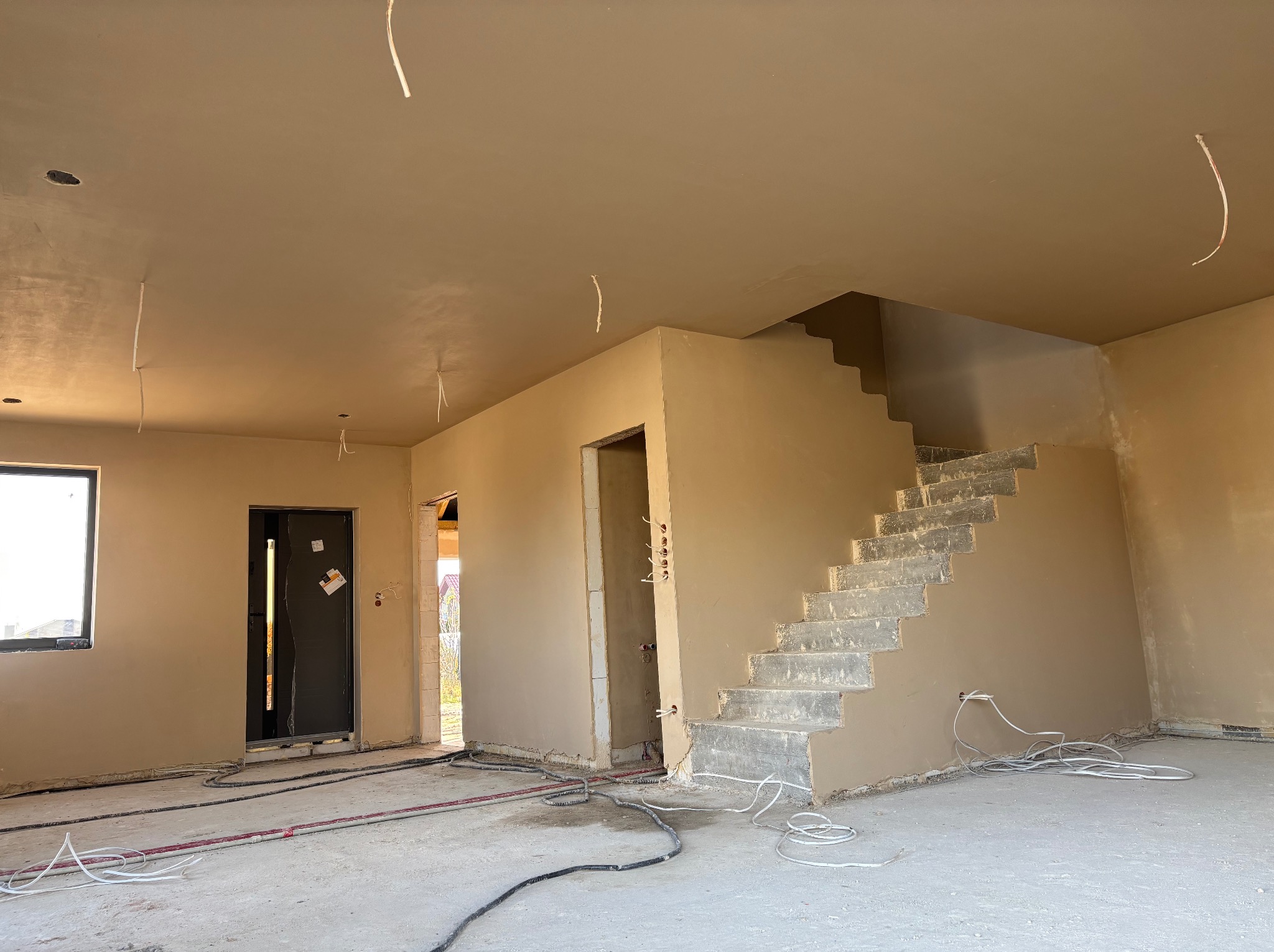 Wnętrze domu po tynkowaniu maszynowym: gładkie ściany w odcieniach beżu, surowe schody betonowe, wystające przewody elektryczne, widoczne otwory drzwiowe i okienne.