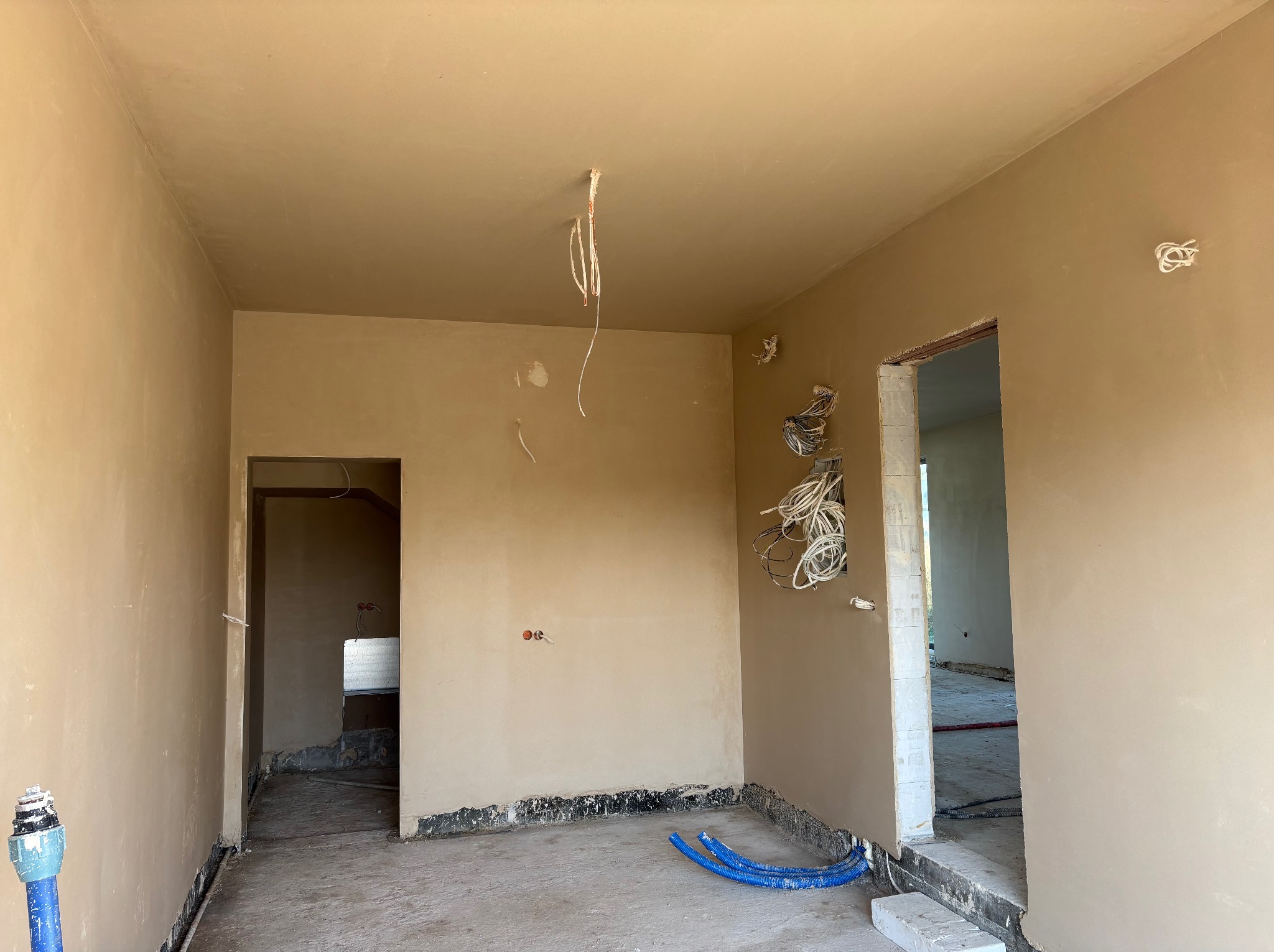 Wnętrze po nałożeniu gładzi gipsowej na ścianach i suficie. Widoczne wystające przewody elektryczne i rury instalacyjne przy podłodze. Przygotowanie do dalszych prac wykończeniowych.