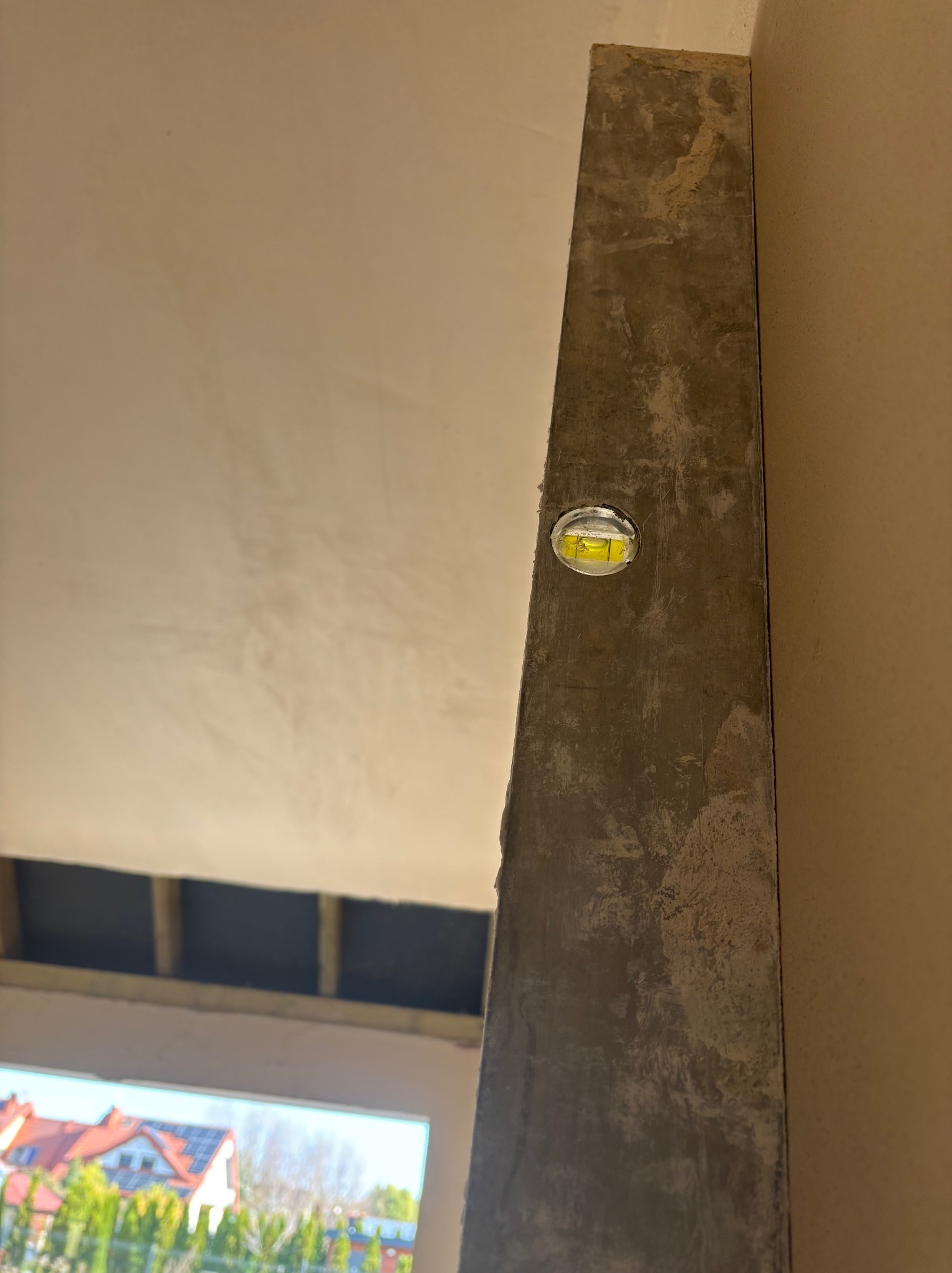 Pionowy profil z poziomnicą w trakcie prac tynkarskich, widok na zewnątrz przez okno. Świeży tynk na ścianie w surowym wnętrzu.