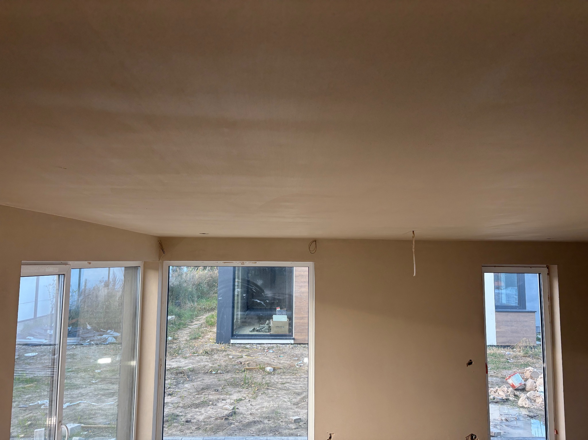 Wnętrze po nałożeniu tynku maszynowego: gładkie ściany i sufit w jasnym odcieniu beżu, widoczne okna z widokiem na zewnątrz, wystające kable elektryczne.