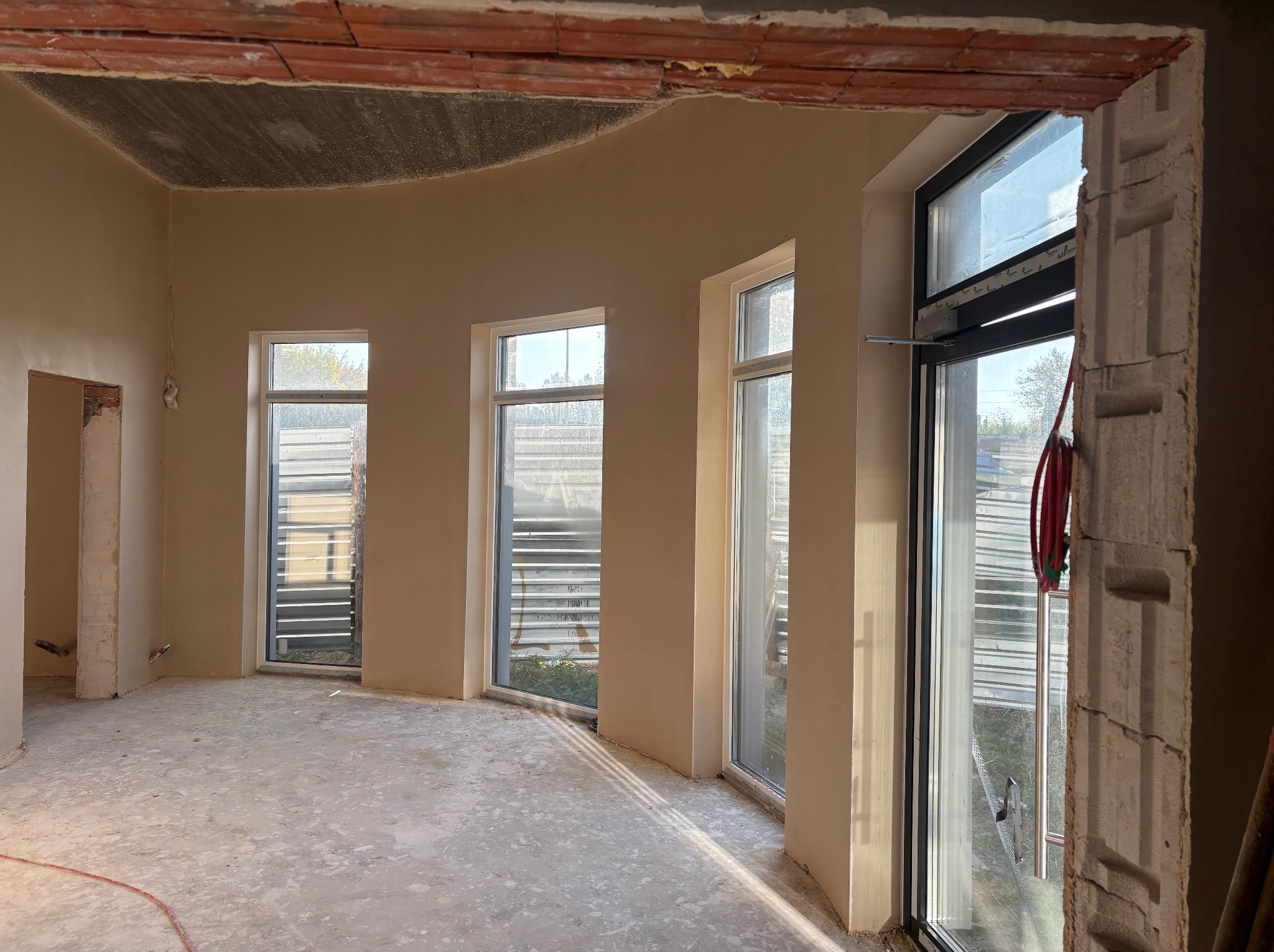 Wnętrze w stanie deweloperskim po nałożeniu gładzi gipsowej na ściany. Widoczne okna, surowy betonowy sufit i fragmenty instalacji elektrycznej.
