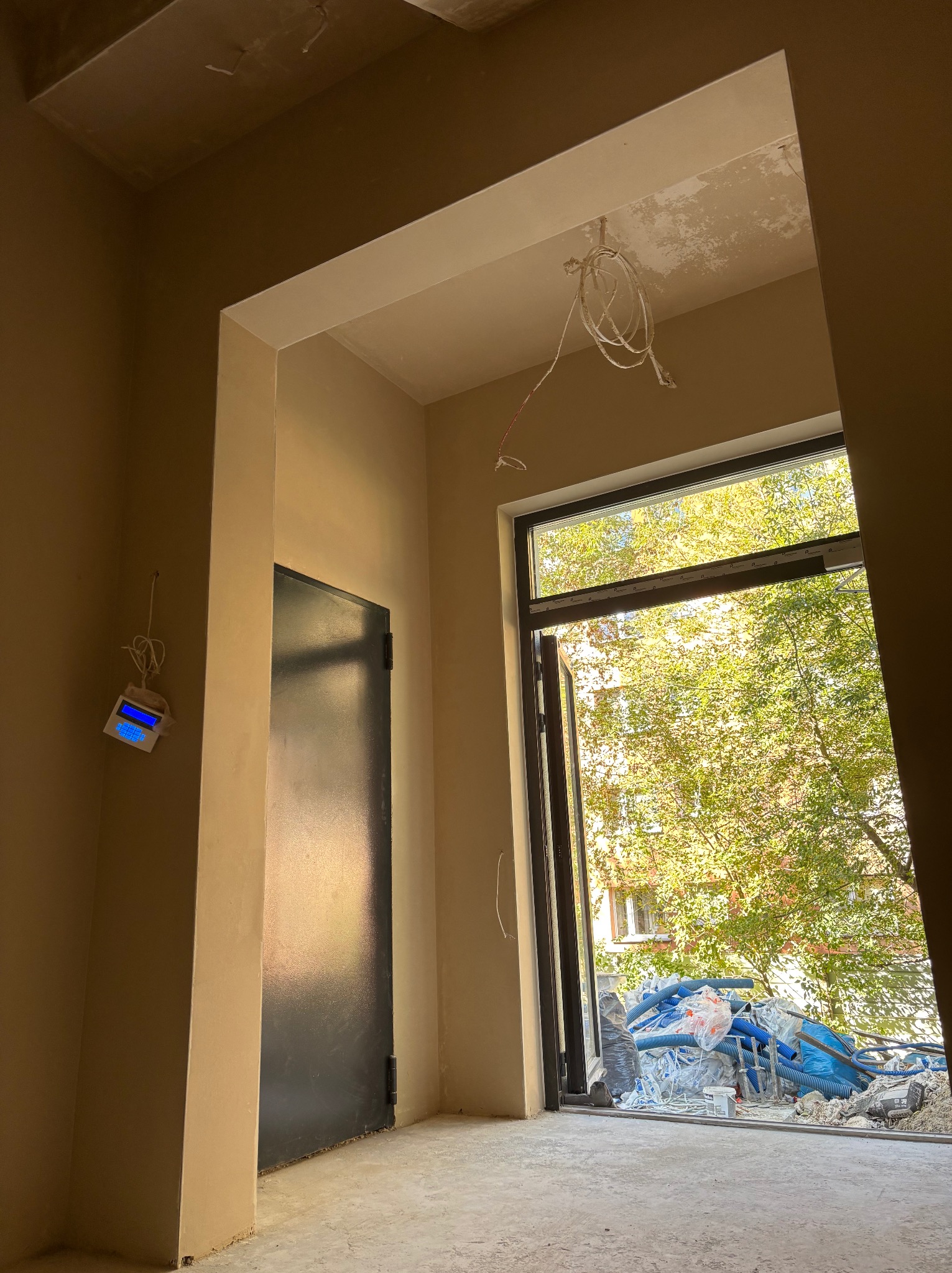Wnętrze w trakcie prac wykończeniowych: świeżo otynkowane ściany w beżowym odcieniu, czarne drzwi i okno z widokiem na zieleń. Na suficie widoczne kable.