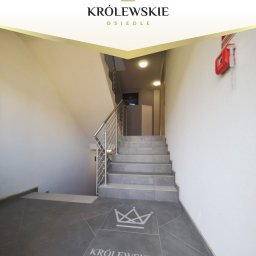 Astral Deweloper - Budowa i Sprzedaż Mieszkań w Stargardzie - Klatka schodowa z logo osiedla w Stargardzie. Szare płytki na podłodze i schodach, metalowe barierki. Na ścianie czerwona skrzynka przeciwpożarowa.