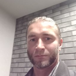 Wiecek - Portret mężczyzny z brodą na tle ściany z szarych cegieł. Ujęcie z góry, oświetlenie mieszane, widoczna część ubrania.