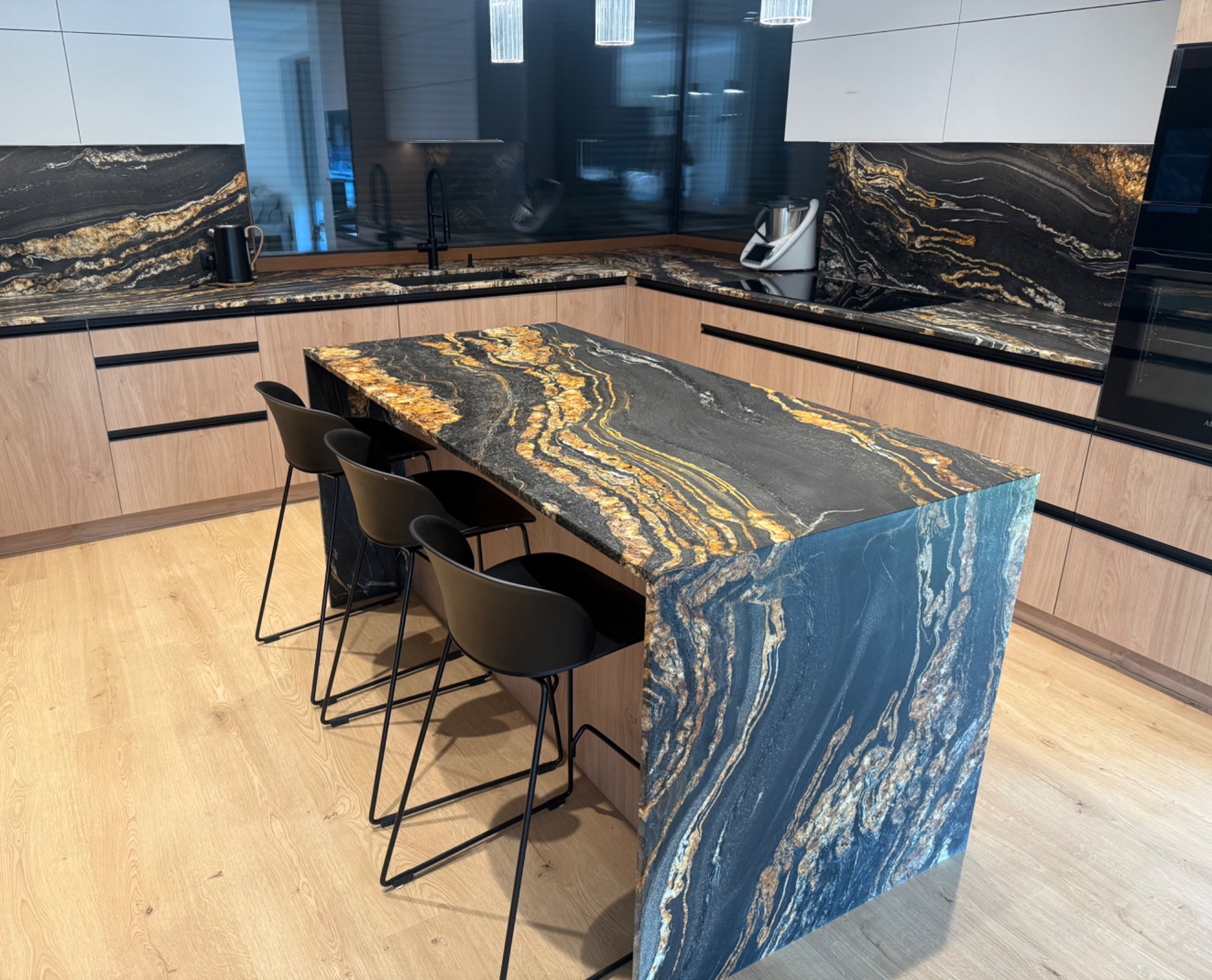 Wnętrze kuchni z wyspą i blatem z czarnego kamienia z wyraźnym, złotym użyleniem. Nowoczesne krzesła barowe i drewniane szafki. Całość w jasnym, naturalnym oświetleniu.