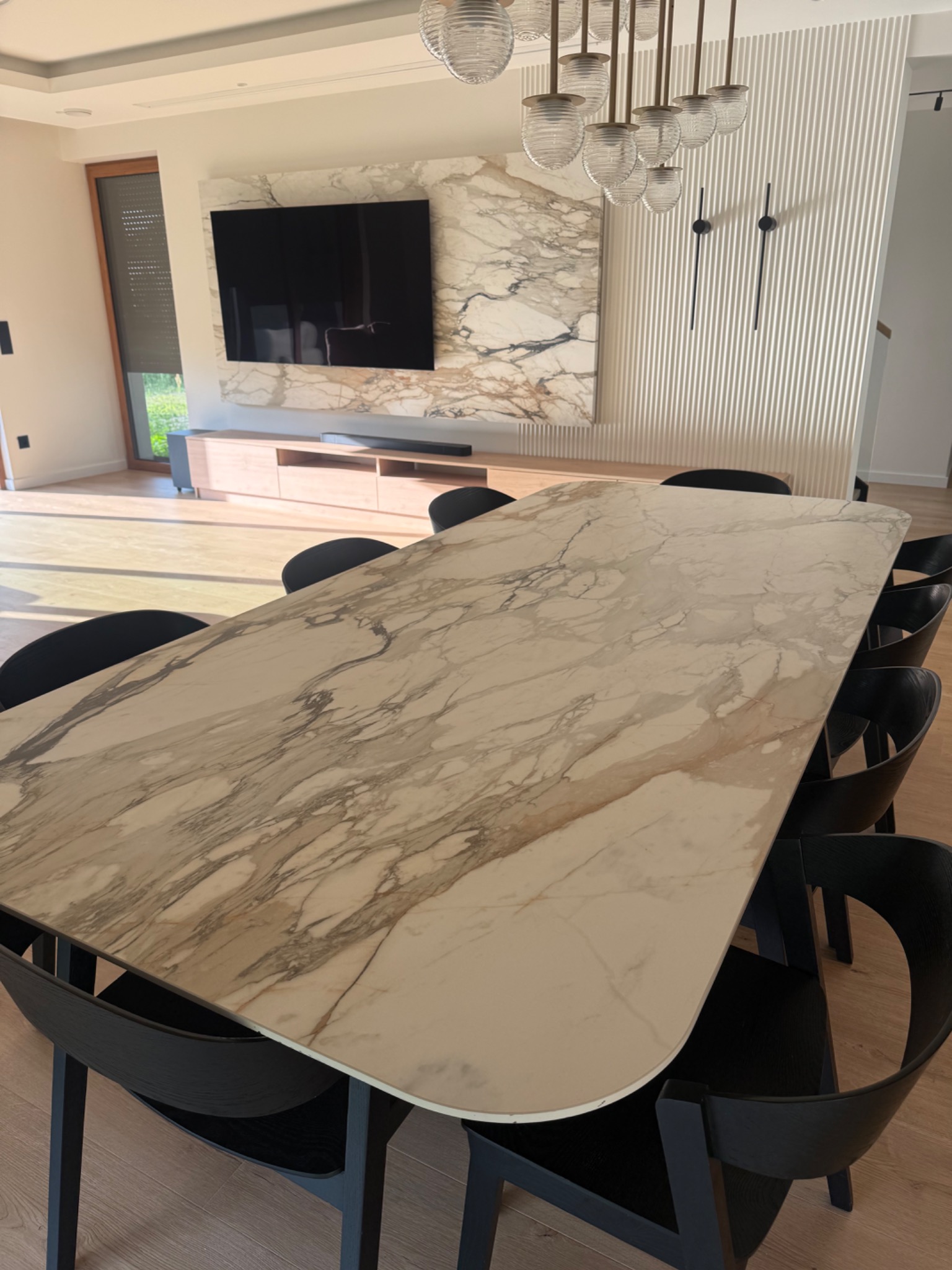 Długi stół z blatem z kamienia naturalnego o marmurkowym wzorze, otoczony czarnymi krzesłami w nowoczesnym wnętrzu z dekoracyjną ścianą i oświetleniem.