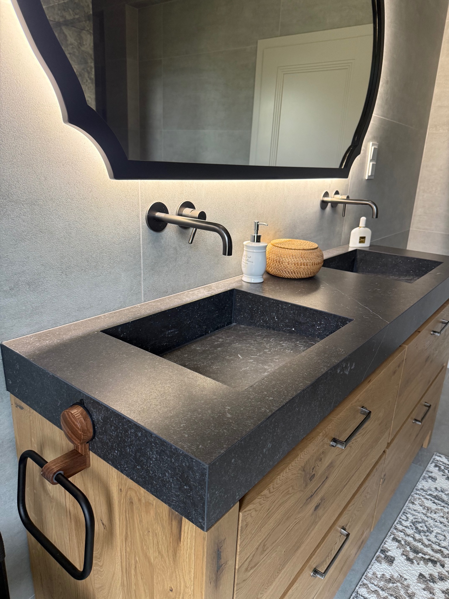 Nowoczesna łazienka z podwójną umywalką i blatem z ciemnego kamienia, drewniana szafka z szufladami, czarne baterie i designerskie lustro z podświetleniem LED.