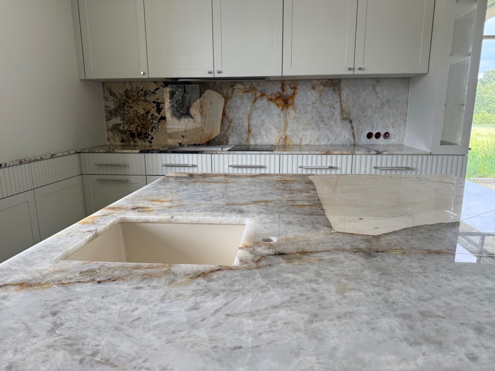 Biały blat kamienny z wyciętym otworem na zlew, widoczny fragment kuchni z białymi szafkami i dekoracyjnym panelem ściennym z kamienia.