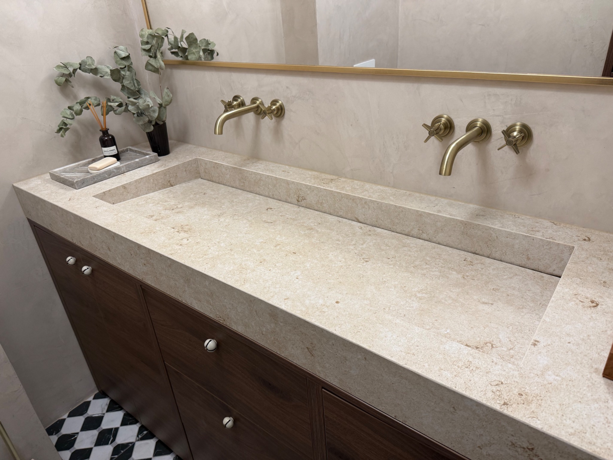 Beżowa, prostokątna umywalka z blatem z konglomeratu, wbudowana w ciemnobrązową szafkę łazienkową. Na blacie dekoracje i złota armatura.