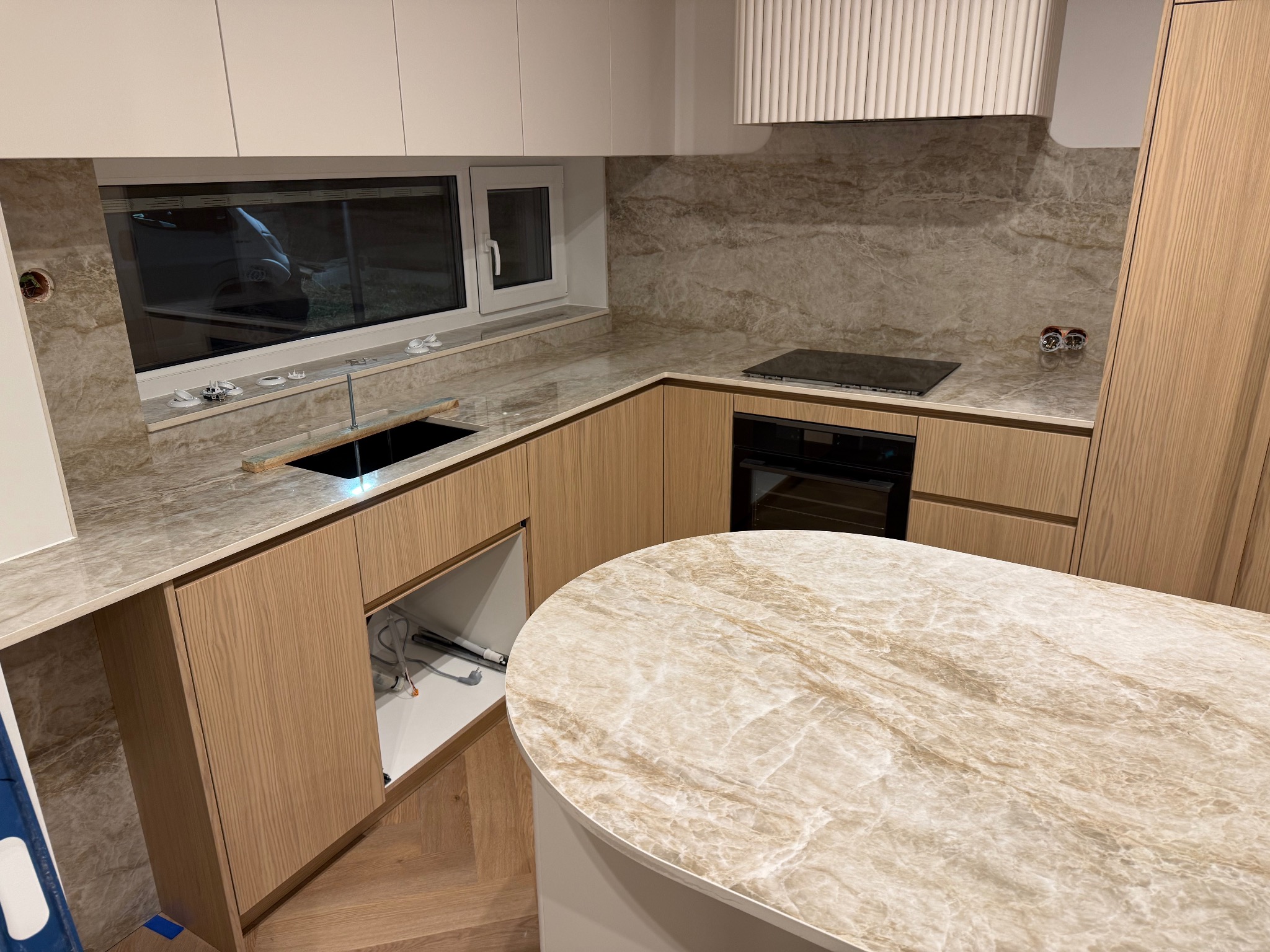 Beżowy blat kamienny w nowoczesnej kuchni z drewnianymi szafkami i oknem. Widoczny fragment wyspy kuchennej z blatem o podobnym wzorze.