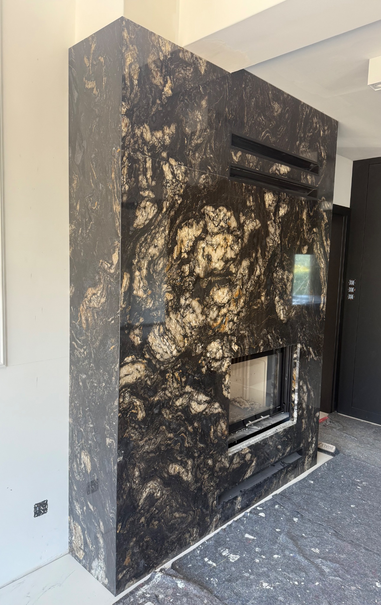 Obudowa kominka z czarnego kamienia z unikalnym wzorem, wkomponowana we wnętrze. Widoczne detale wykończenia i otwory wentylacyjne. Podłoga zabezpieczona folią.