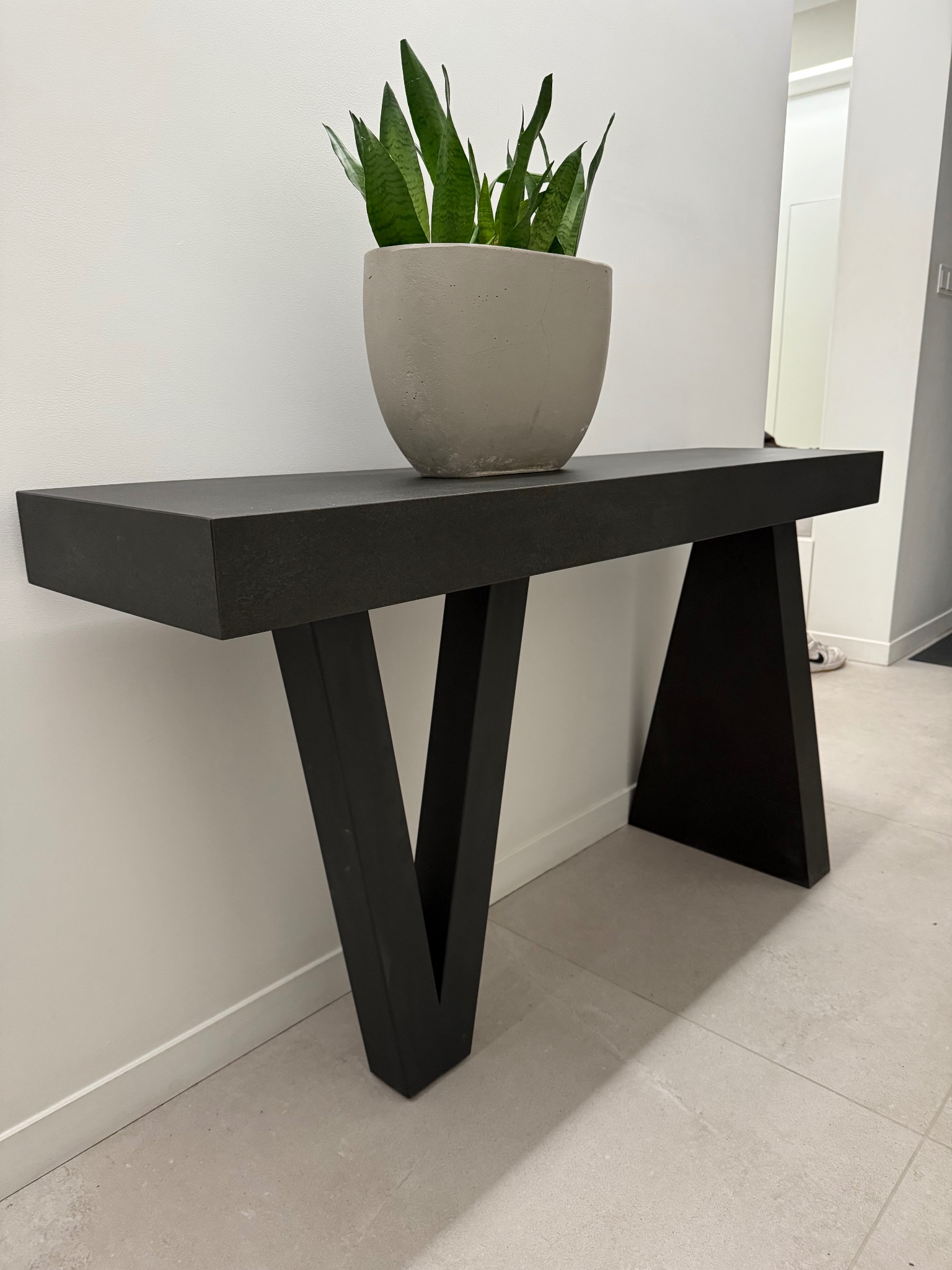Nowoczesny blat z ciemnego kamienia na geometrycznych nogach, zwieńczony donicą z zieloną rośliną. Minimalistyczne wnętrze z jasnymi ścianami i podłogą.