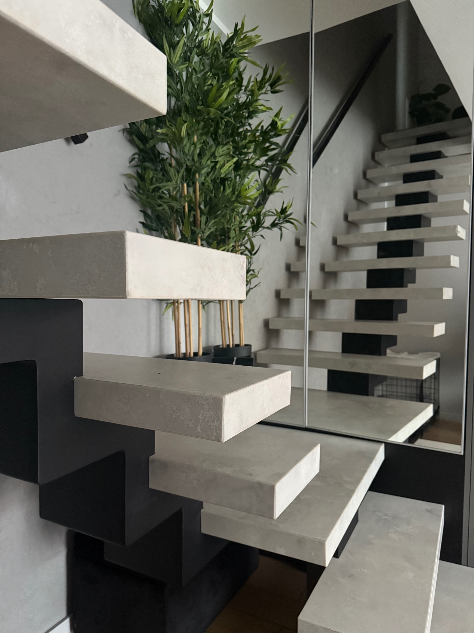 Nowoczesne schody wspornikowe z betonu architektonicznego, z minimalistyczną konstrukcją i rośliną doniczkową w tle, odbijające się w lustrze.