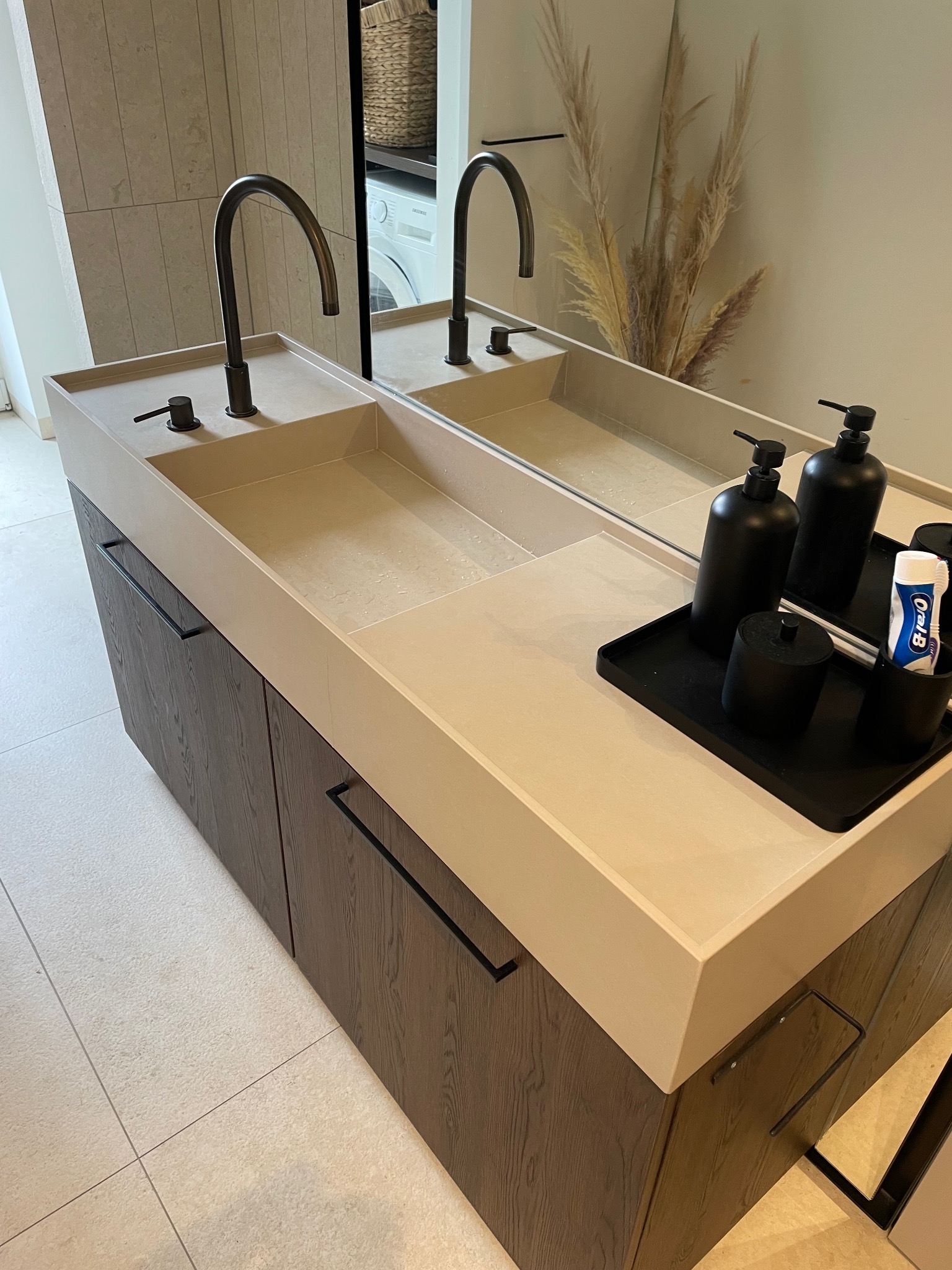 Beżowa umywalka z szafką z ciemnego drewna, czarne baterie i akcesoria łazienkowe. W tle lustro odbijające pralkę i dekoracyjne trawy.