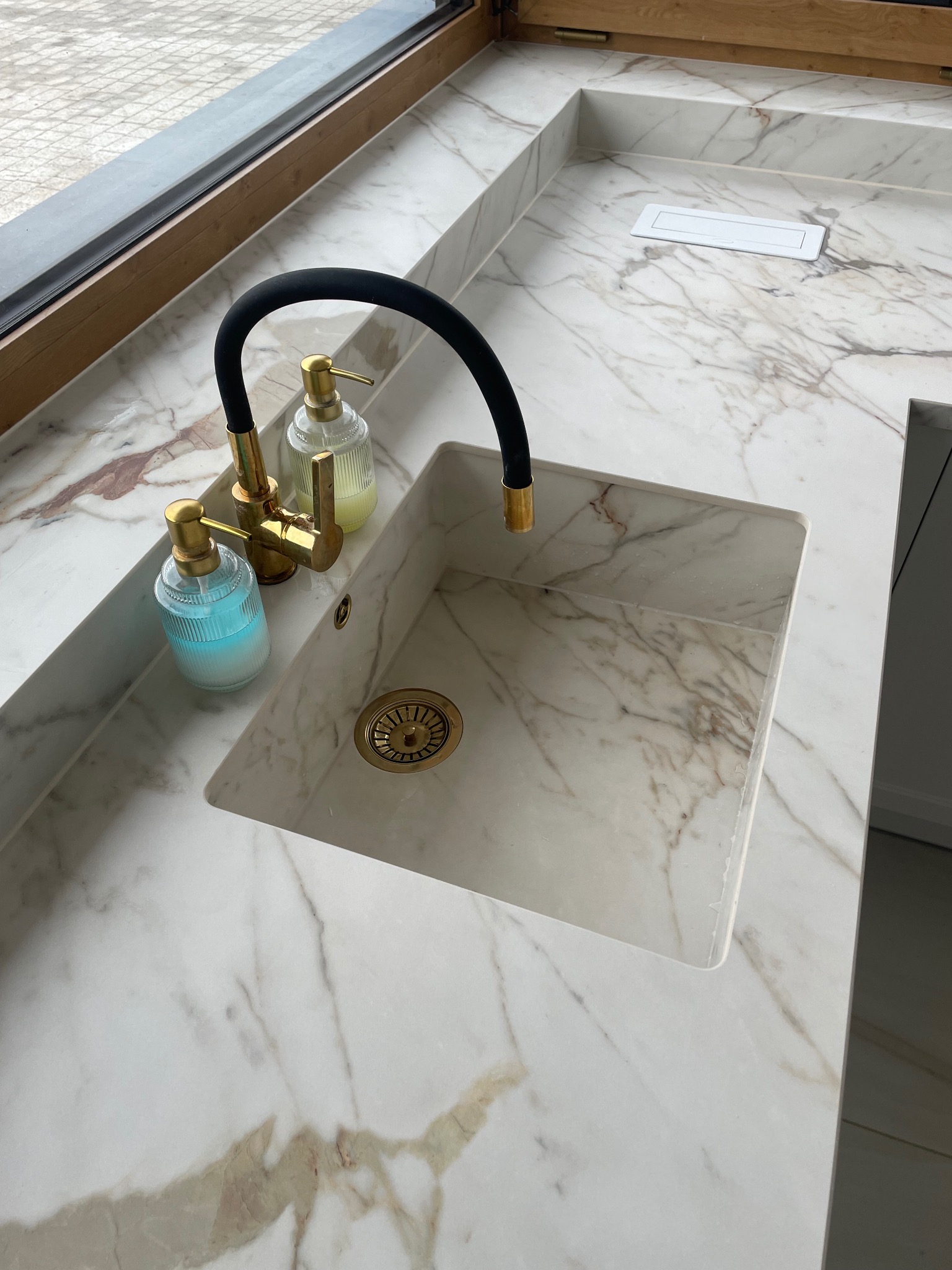 Marmurowy blat kuchenny z wbudowanym zlewem, złotą armaturą i dozownikami mydła. Widoczny fragment okna i gniazdko elektryczne w blacie. Ujęcie z góry.