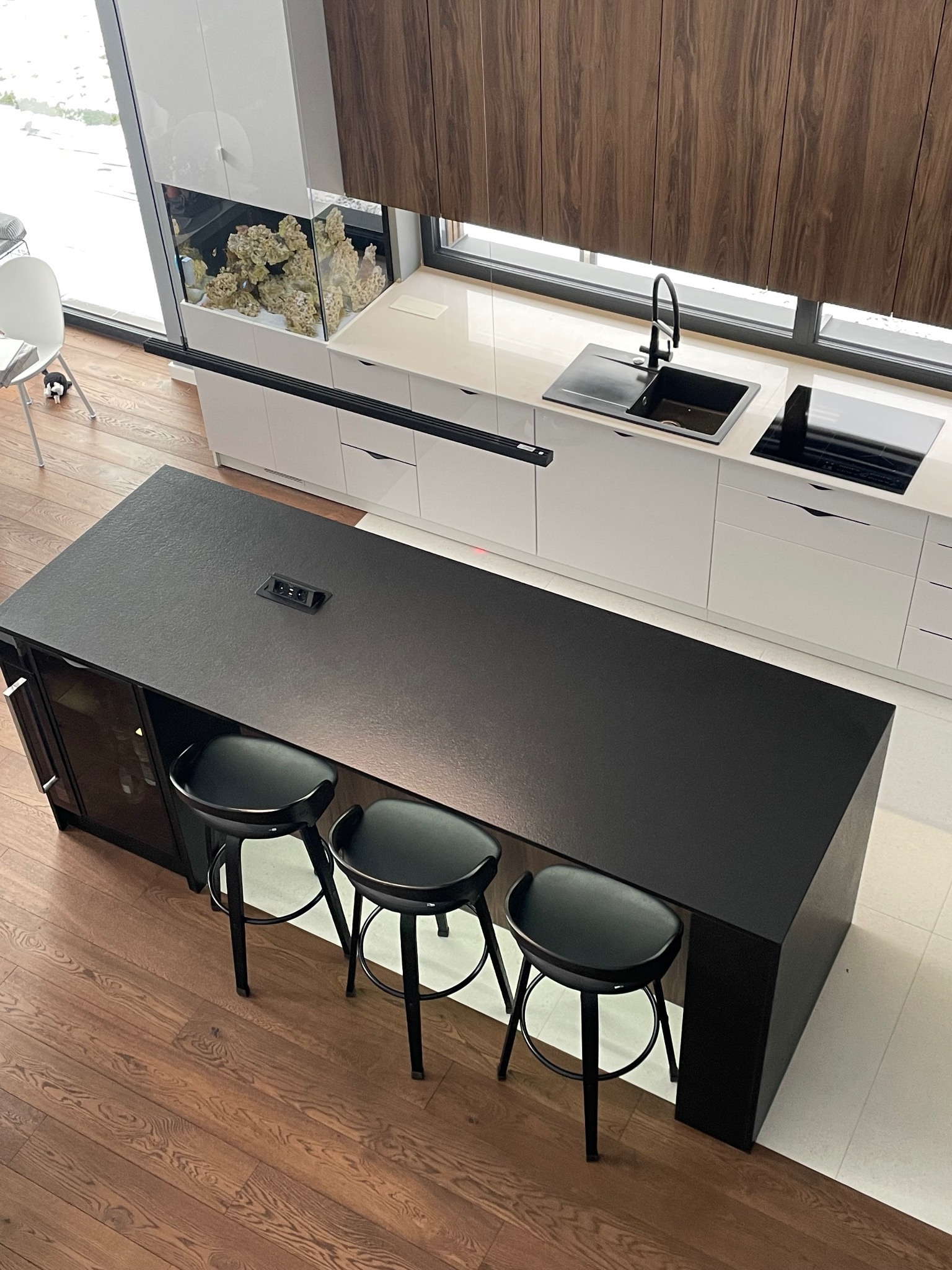 Nowoczesna kuchnia z czarnym blatem kamiennym na wyspie, trzema hokerami i akwarium w zabudowie. Minimalistyczny design, drewniane akcenty, widok z góry.