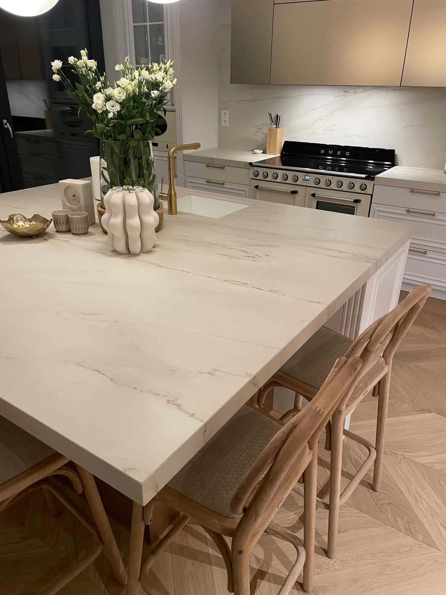 Jasny blat kamienny w kuchni z drewnianymi krzesłami barowymi, kwiatami i dekoracjami. Widoczny fragment kuchenki SMEG i parkiet w jodełkę.