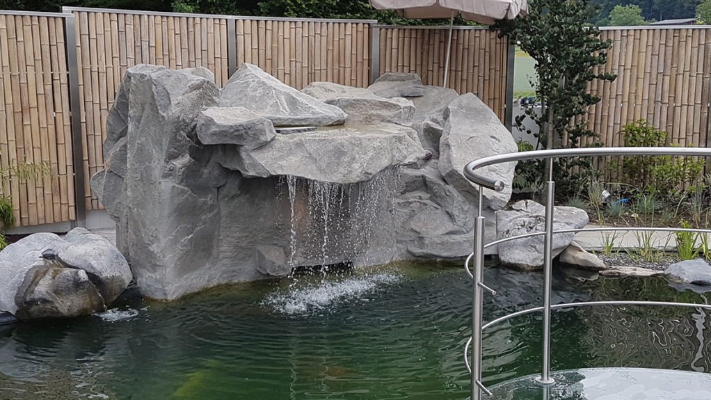 Oczko wodne z kaskadą wodną, otoczone kamieniami i bambusowym ogrodzeniem. W tle widoczna zieleń ogrodu i fragment parasola.