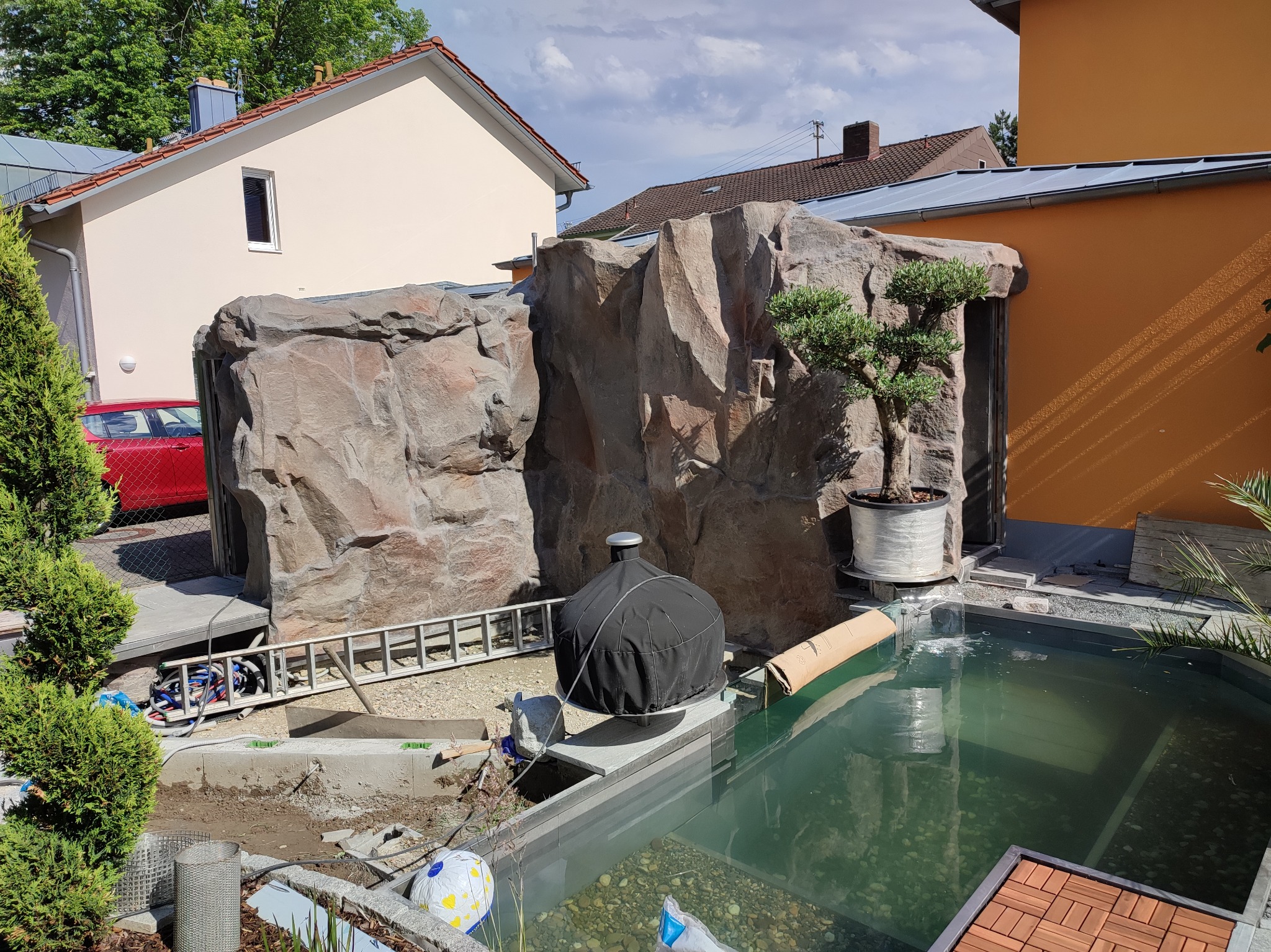 Nowoczesny basen ogrodowy z dekoracyjnymi skałami i drzewkiem bonsai, w trakcie budowy, z widocznymi narzędziami i elementami instalacji.