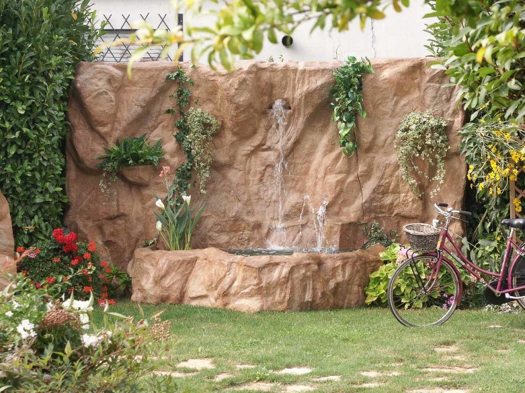 Ogród z kaskadowym, sztucznym wodospadem z kamienia, otoczonym zielenią i kwiatami. Różowy rower z koszykiem dodaje uroku.