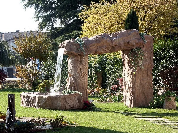 Ogród z dekoracyjną, kamienną fontanną w formie portalu, z której spływa woda do małego zbiornika. Otoczenie zdobi trawa i roślinność.
