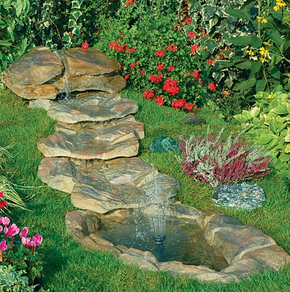 Kaskada wodna z kamieni w ogrodzie, otoczona zielenią i kwitnącymi roślinami. Woda spływa po kamieniach do małego zbiornika.